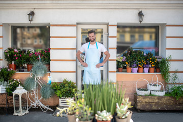 Young bearded male standing at the door of flower shop with hands on his hips