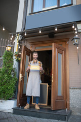 bearded waiter in grey apron standing at the entrance, holding open sign, smiling, greeting