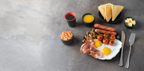 Typical English breakfast on dark concrete background. Fried eggs, sausages, bacon, beans, mushrooms, toasts and butter, coffee and orange juice. Copy space