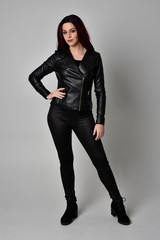 Full length portrait of  girl with red hair wearing black leather jacket, pants and boots. Standing pose, isolated against a grey studio background.