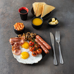 Typical English breakfast on dark concrete background. Fried eggs, sausages, bacon, beans, mushrooms, toasts and butter, coffee and orange juice. Copy space  