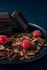Chocolate pieces with chocolate shavings and raspberry in black plate on dark wooden background