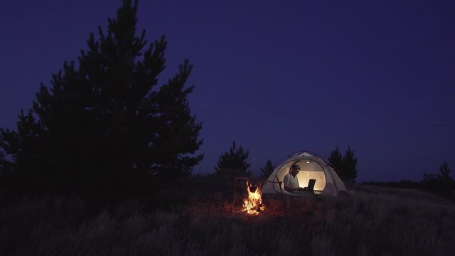 Man Works behind a Laptop in a tent near a fire at night. Freelancer Remote Work Concept. 4K