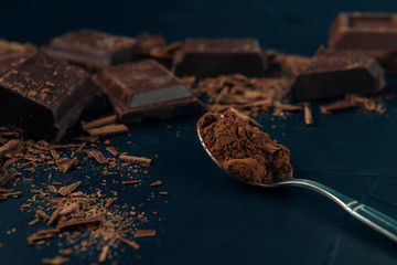 Pieces of chocolate, chocolate shavings and spoon with cocoa powder on dark blue textured background.