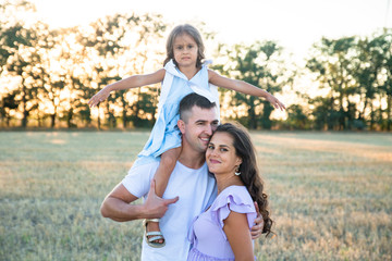 happy young husband, pregnant wife and a little girl on the father's shoulders on the field, wind turbines on the background