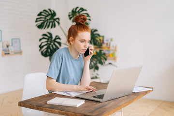 Serious redhead young businesswoman working on laptop computer at the desk and talking on mobile phone in light cozy room at the home office. Concept of distance job on Internet in self isolation.