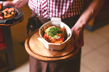 Italian lasagna in a white bowl. Waiter is holding a plate with traditional Italian food, tomato meat lasagna.