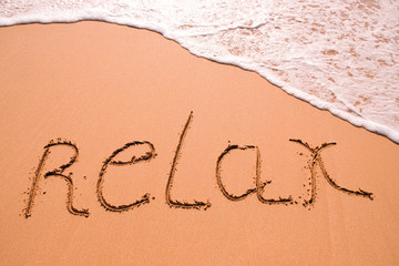 Word Relax hand written in the sand with a sea wave. Close up sand texture on beach in summer.