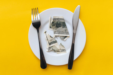 The dollar bill lies on a white plate, pierced with a fork near the knife on a yellow cotton...