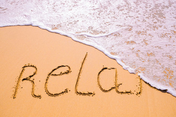 Word Relax hand written in the sand with a sea wave. Close up sand texture on beach in summer.