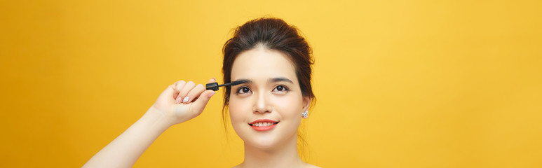 Fototapeta premium Closeup portrait of attractive young woman putting some mascara onto her eyelashes with make up brush over yellow background