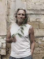 Obraz premium Smoking gray-haired man is standing at wall of an old abandoned estate and holds branch of cannabis in his hand. Vintage effect.