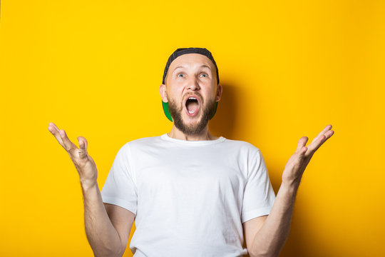 Rejoices, Delighted Young Man In Baseball Cap Holding Palms Up On Yellow Background.