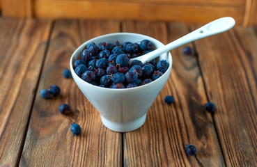 Close up view of a white cup full of fresh shadberry with ceramic tea spoon on old grunge wooden table. A few berries are scattered on the table. Selective focus.