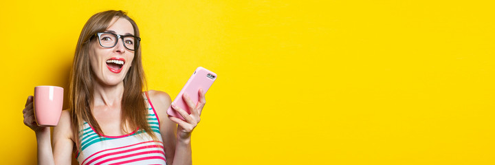 Young woman rejoices with a cup of coffee and with a phone on a yellow background.