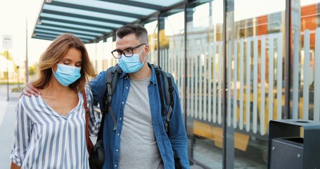 Caucasian handsome man and beautiful woman walking outdoors, talking and hugging. Couple of travellers in medical masks strolling at bus stop ot train station. Pandemic travelling. Covid-19 concept.