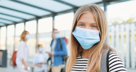 Fototapeta na wymiar Portrait of pretty Caucasian blonde cute teen girl in medical mask standing at bus stop outdoor and looking at camera. Zooming in. Parents with small brother and suitcases on blurred background.