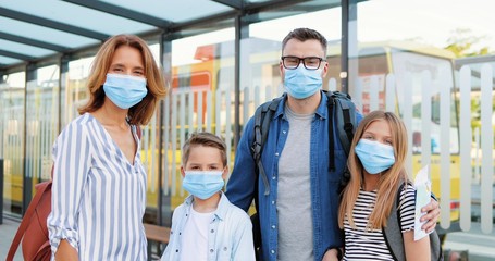 Portrait of Caucasian happy family in medical masks and with suitcases standing outdoor at bus sop or train station. Parents with son and daughter travelling during pandemic. Zooming in.