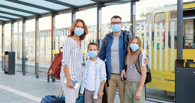 Portrait of Caucasian happy family in medical masks and with suitcases standing outdoor at bus sop or train station. Parents with son and daughter travelling during pandemic. Zooming in.