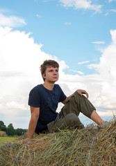 Naklejka na ściany i meble A handsome young man is sitting on a haystack against a blue cloudy sky.
