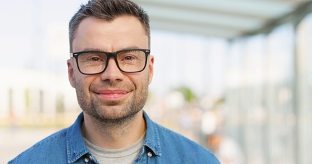 Portrait of handsome Caucasian man in glasses looking straight and smiling to camera outdoor. Close up of attractive happy male in eyeglasses at bus stop on summer sunny day. Zooming in face.