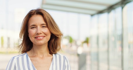 Portrait shot of beautiful Caucasian woman with fair hair looking at side, turning face to camera and smiling outdoors on sunny summer day. Happy female at street. Close up of cheerful lady.