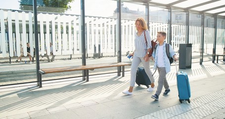 Caucasian young beautiful happy mother and cute small son walking the street and carrying suitcases on wheels at bus stop. Woman and little boy smiling, talking and having vacations. Train station.