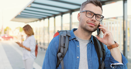 Caucasian handsome young man in glasses and with backpack standing at bus stop and talking on mobile phone. Male in eyeglasses speaking on cellphone at train station. Telephone call.