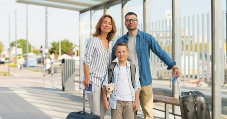 Fototapeta premium Portrait of Caucasian happy family with suitcases standing outdoors at bus sop or train station. Parents with son travelling in summer. Vacations of child with mother and father Departure for holidays