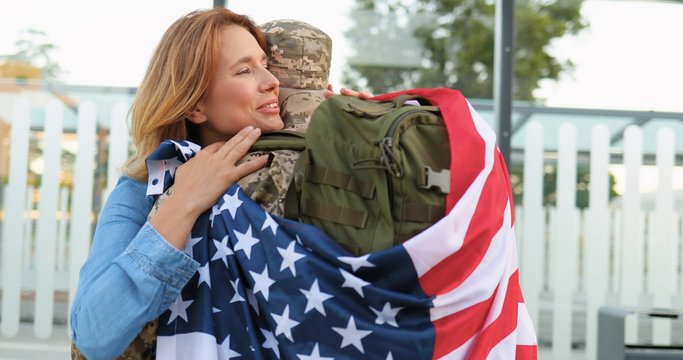 Happy young Caucasian couple meeting and hugging at street. Handsome male soldier in uniform coming back from army and meeting with beautiful wife with USA flag. War service returning.