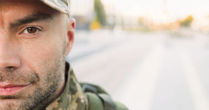 Portrait of Caucasian handsome young man soldier in cap with backpack looking confident at street. Close up of half face of male militarian with wise brave face outdoor at bus stop. Military uniform.