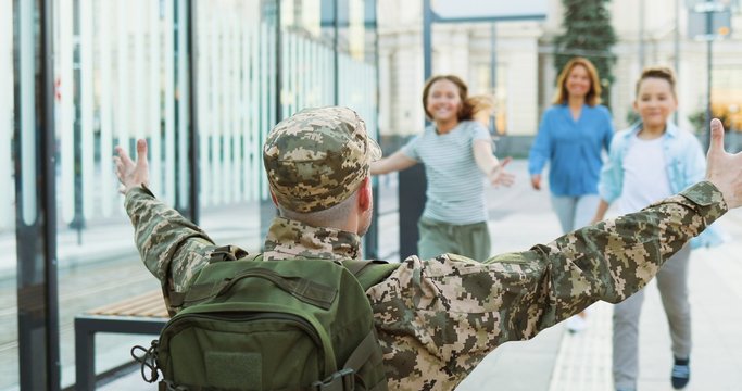 Rear of soldier in military uniform standing at train station with arms open wide and kids with wife running to hug him. Returning from army war service. Father officer meeting son, daughter and woman