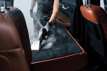 Uses steam cleaner. Modern black automobile is in service by woman inside of car wash station