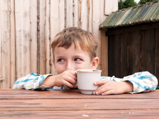 Boy is sitting at wooden table with cup of tea and looking into distance.