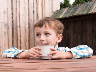 Boy is sitting at wooden table with cup of tea and looking into distance.