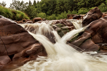 Long exposure image of a small creek flowing water through brown stones. Green trees in the...