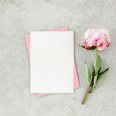Mockup invitation, blank paper greeting card, pink envelope and peonies on gray stone table. Flower background. Flat lay, top view.