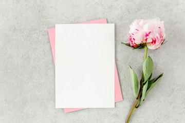 Fototapeta premium Mockup invitation, blank paper greeting card, pink envelope and peonies on gray stone table. Flower background. Flat lay, top view.