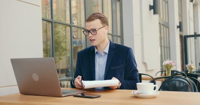 A businessman is indignantly discussing business issues at a meeting via a video call on a laptop. He is sitting in a cafe and on the table a cup of tea. 4K