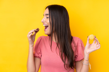 Young brunette woman over isolated yellow background holding colorful French macarons and eating it