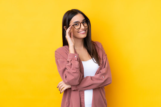 Young Brunette Woman Over Isolated Yellow Background With Glasses And Happy