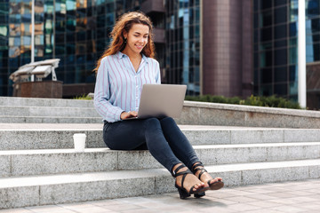 Smiling businesswoman working at laptop outdoors in front of business center sitting on steps
