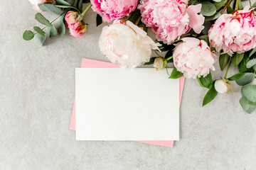 Obraz na płótnie Canvas Mockup invitation, blank paper greeting card, pink envelope and peonies on gray stone table. Flower background. Flat lay, top view.