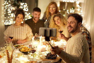 holidays, celebration and people concept - happy friends drinking non-alcoholic red wine and taking picture with smartphone on selfie stick at home christmas dinner party