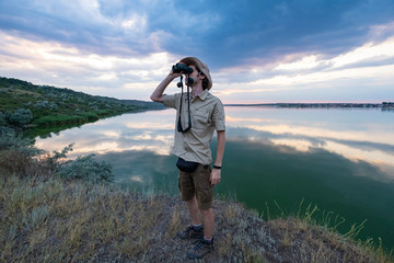 Young male traveler or 
scientist with binocular and photo camera outdoors near river. 