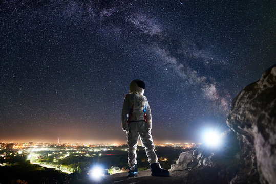 Mission specialist astronaut standing on rocky hill and looking at beautiful starry sky with milky way. Space traveler wearing white space suit and helmet. Concept of human space exploration. - Powered by Adobe