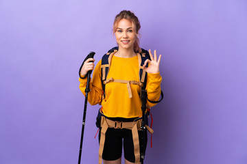 Teenager girl with backpack and trekking poles over isolated purple background showing ok sign with two hands