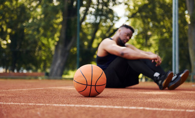 Tired african american man sits on the ground with ball on the court outdoors. Takes a break