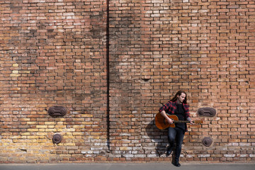 Young male with long hair play on acoustic guitar near brown wall 