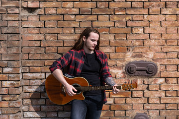Young male with long hair play on acoustic guitar near brown wall 